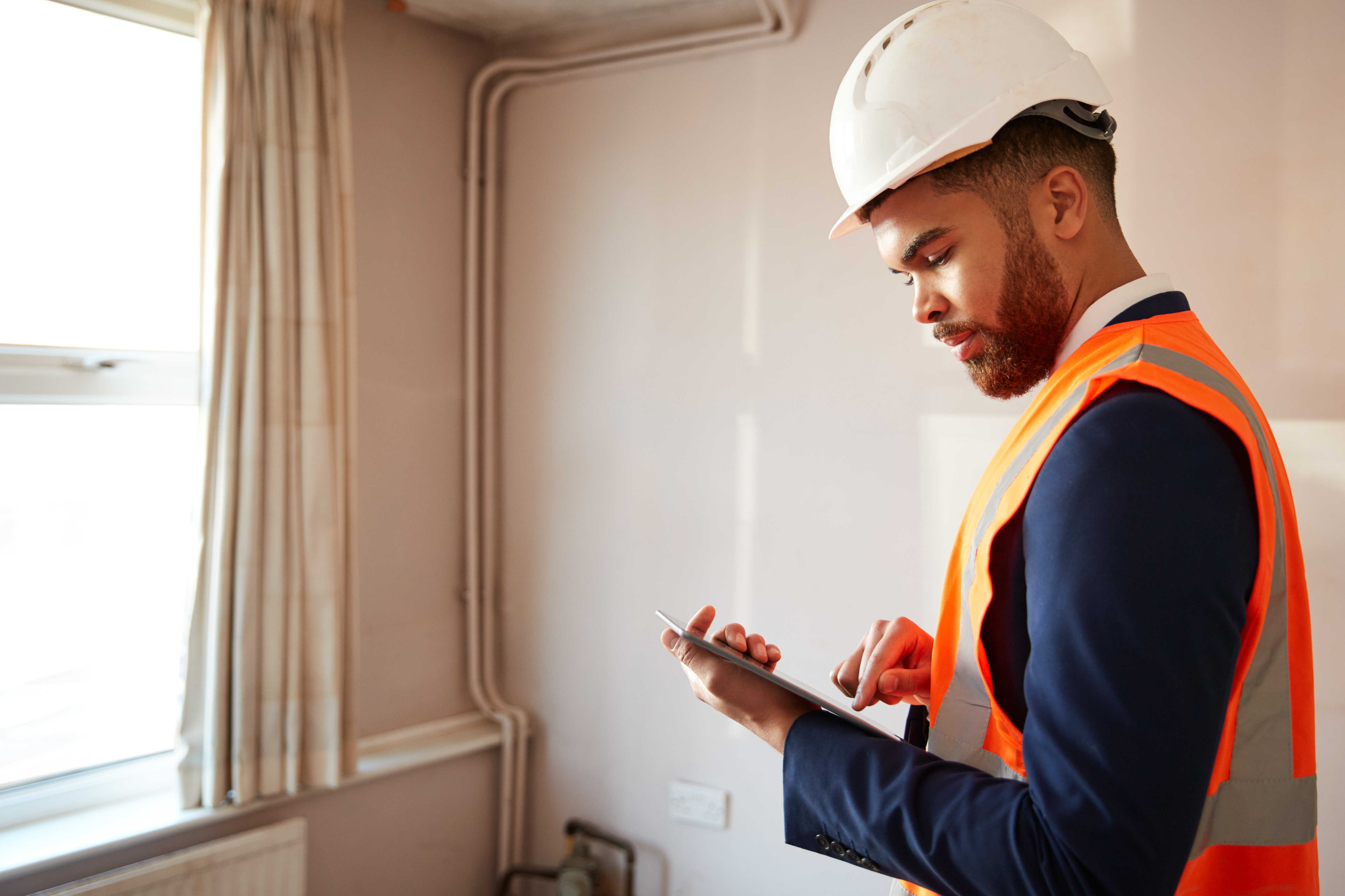 A man wearing an orange vest and hard hat is focused on using a tablet at a home for survey.